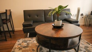 Ein runder Holztisch mit einer Zimmerpflanze in der Mitte, ein gemustertes Teppich, eine schwarze Couch und ein kleiner Beistelltisch mit einer Lampe.
