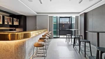 A modern bar area with a long counter and multiple stools, a kitchen visible through a glass partition, and a seating area with tables and chairs.