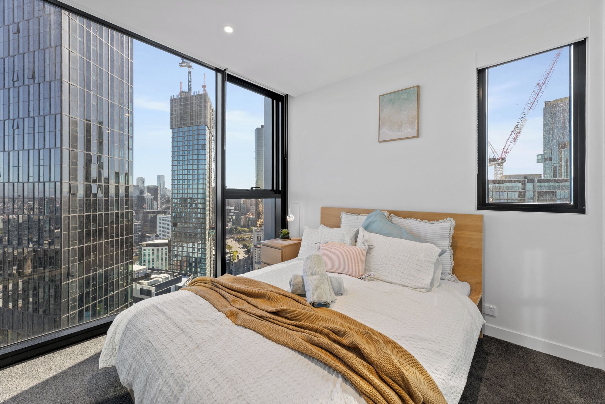 A bedroom with a large bed, a wooden headboard, and a view of a cityscape through floor-to-ceiling windows.