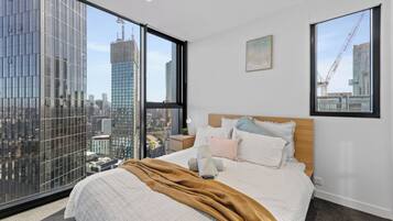 A bedroom with a large bed, a wooden headboard, and a view of a cityscape through floor-to-ceiling windows.