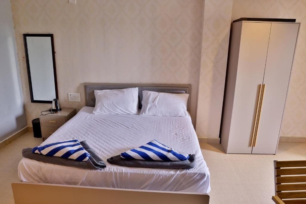 A neatly made bed with white linens and striped pillows, a bedside table with a lamp, a wardrobe, and a mirror on the wall.