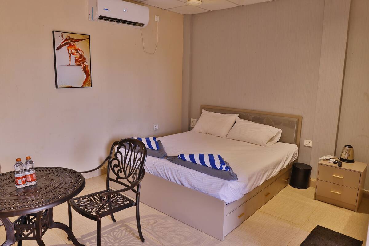 A bedroom with a bed, a chair, a small table with bottles, and a trash bin.