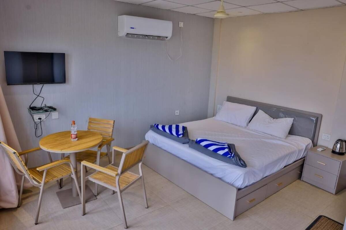 A hotel room with a bed, a TV, an air conditioner, a table with chairs, and a bedside table with a kettle.