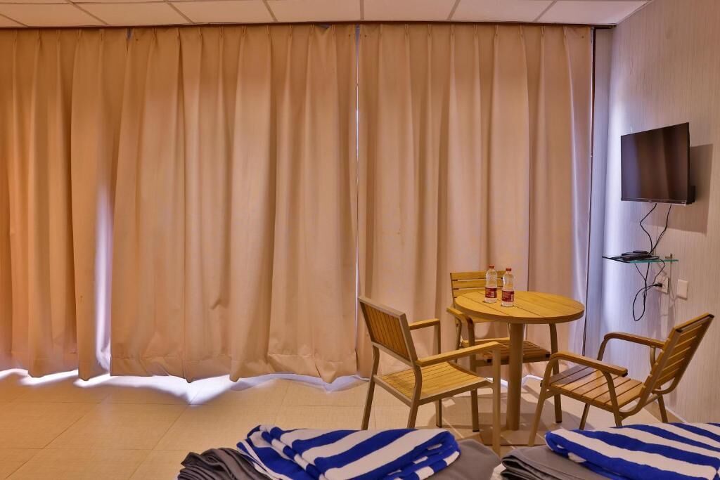 Una habitación con cama, una mesa redonda con sillas, un televisor montado en la pared y una ventana con cortinas.