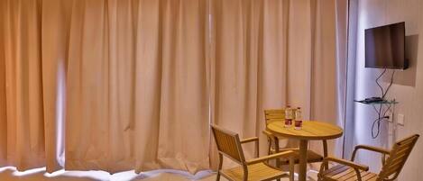 Una habitación con cama, una mesa redonda con sillas, un televisor montado en la pared y una ventana con cortinas.