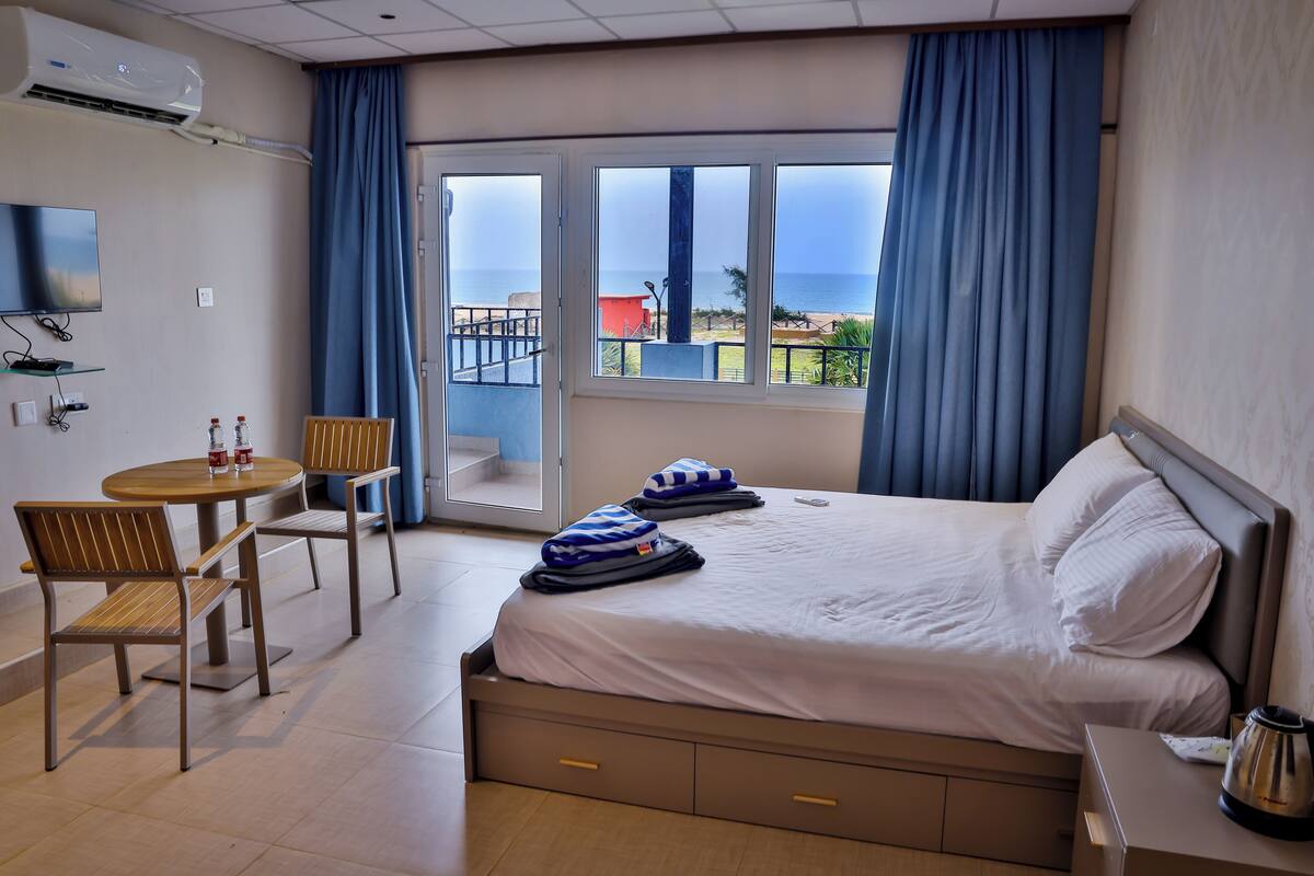 A hotel room with a bed, a desk with a chair, a TV, and a view of the sea.