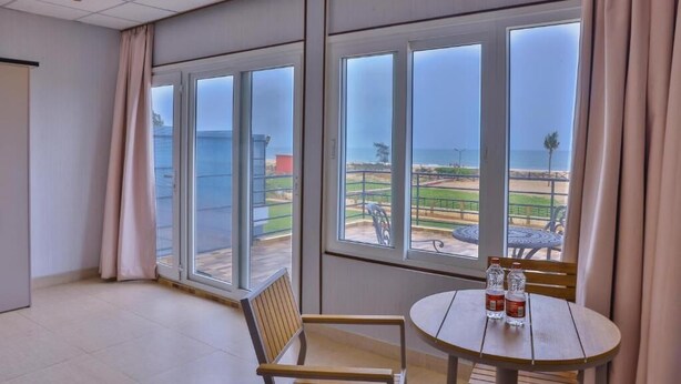 Een kamer met grote ramen, een ronde tafel met stoelen en uitzicht op een strand en een park.