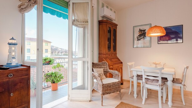 A room with a dining table, chairs, a wicker chair, a cabinet, a potted plant, and a view of buildings through a window. - Stylish flat balcony near Rapallo Castle (Rapallo)
