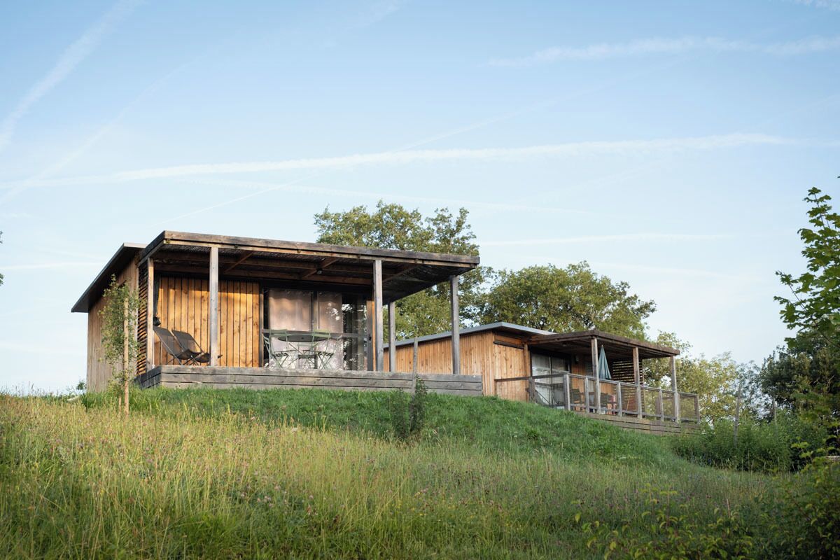A wooden cabin with a porch and a covered outdoor seating area, surrounded by natural landscape.