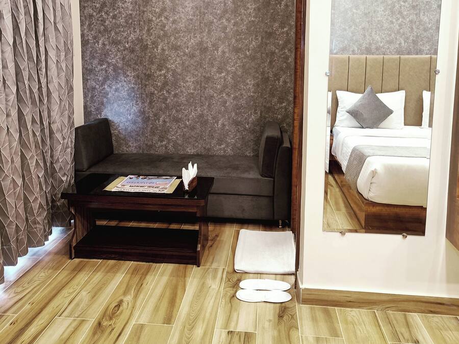 A modern hotel room with a wooden floor, a dark sofa, a small coffee table, and a large mirror reflecting a bed with white bedding and a gray pillow.