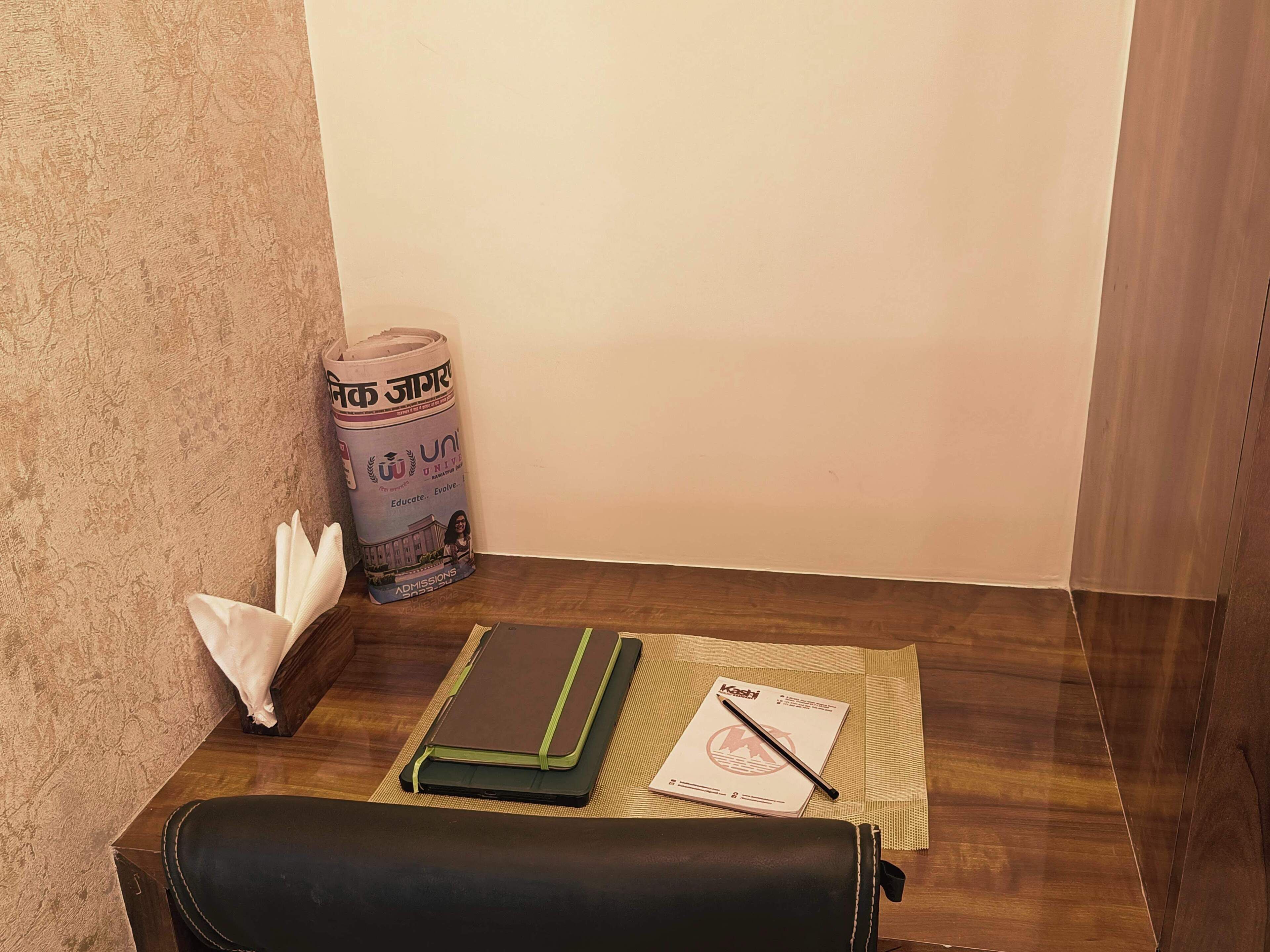 A corner of a room with a wooden floor, a wall with a textured pattern, a rolled-up newspaper, a black leather bag, a green notebook, and a pen on a woven mat.