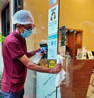 A person in a red shirt and hairnet sanitizing a hand with a spray bottle and a cloth at a reception area with a sign about COVID-19 prevention.