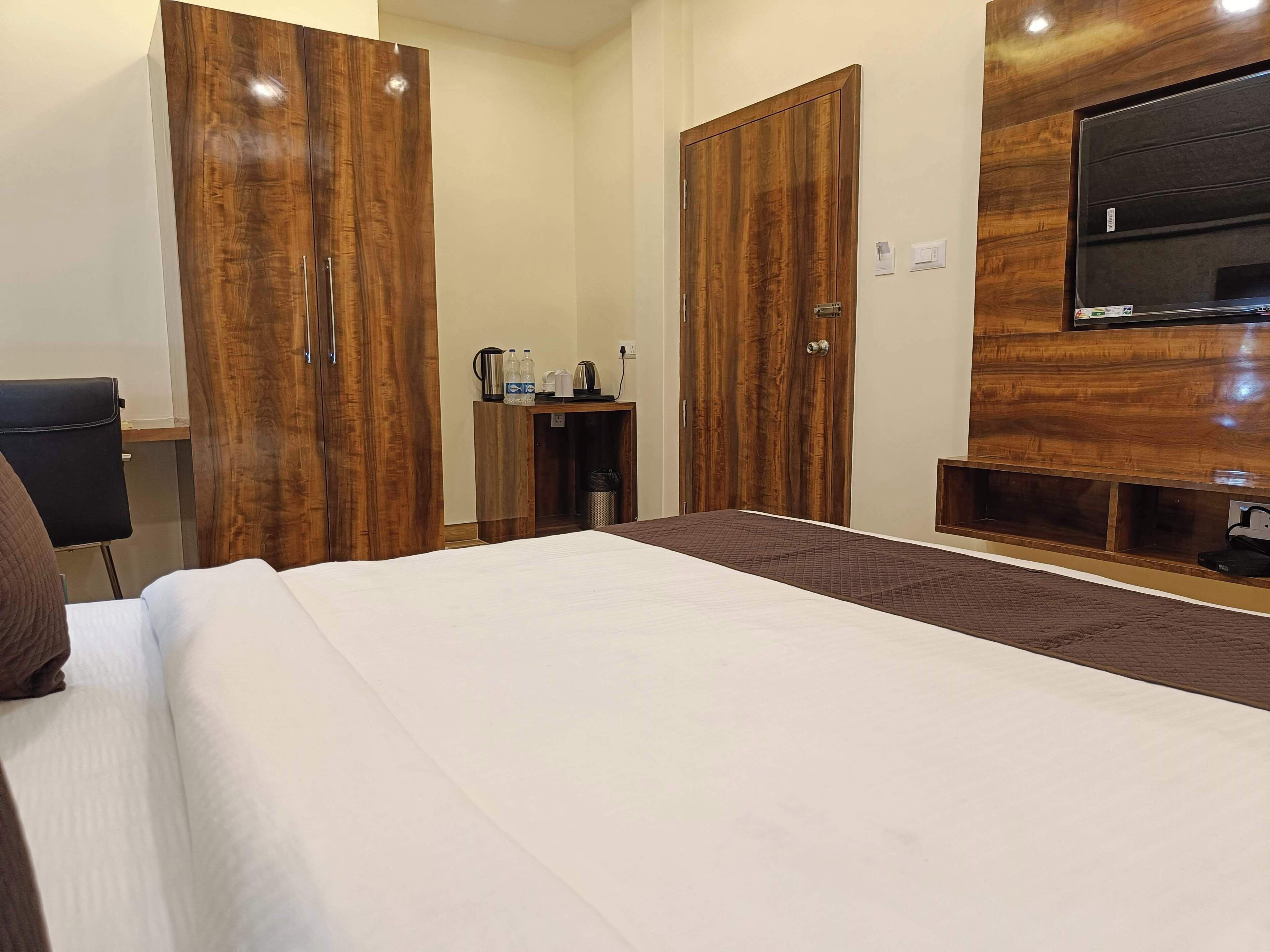 A hotel room with a large bed, wooden wardrobes, a desk with a chair, a television mounted on the wall, and a small table with a kettle and bottles.