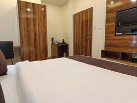 Habitación de hotel con una cama grande, armarios de madera, un escritorio con silla, un televisor montado en la pared y una mesita con tetera y botellas.