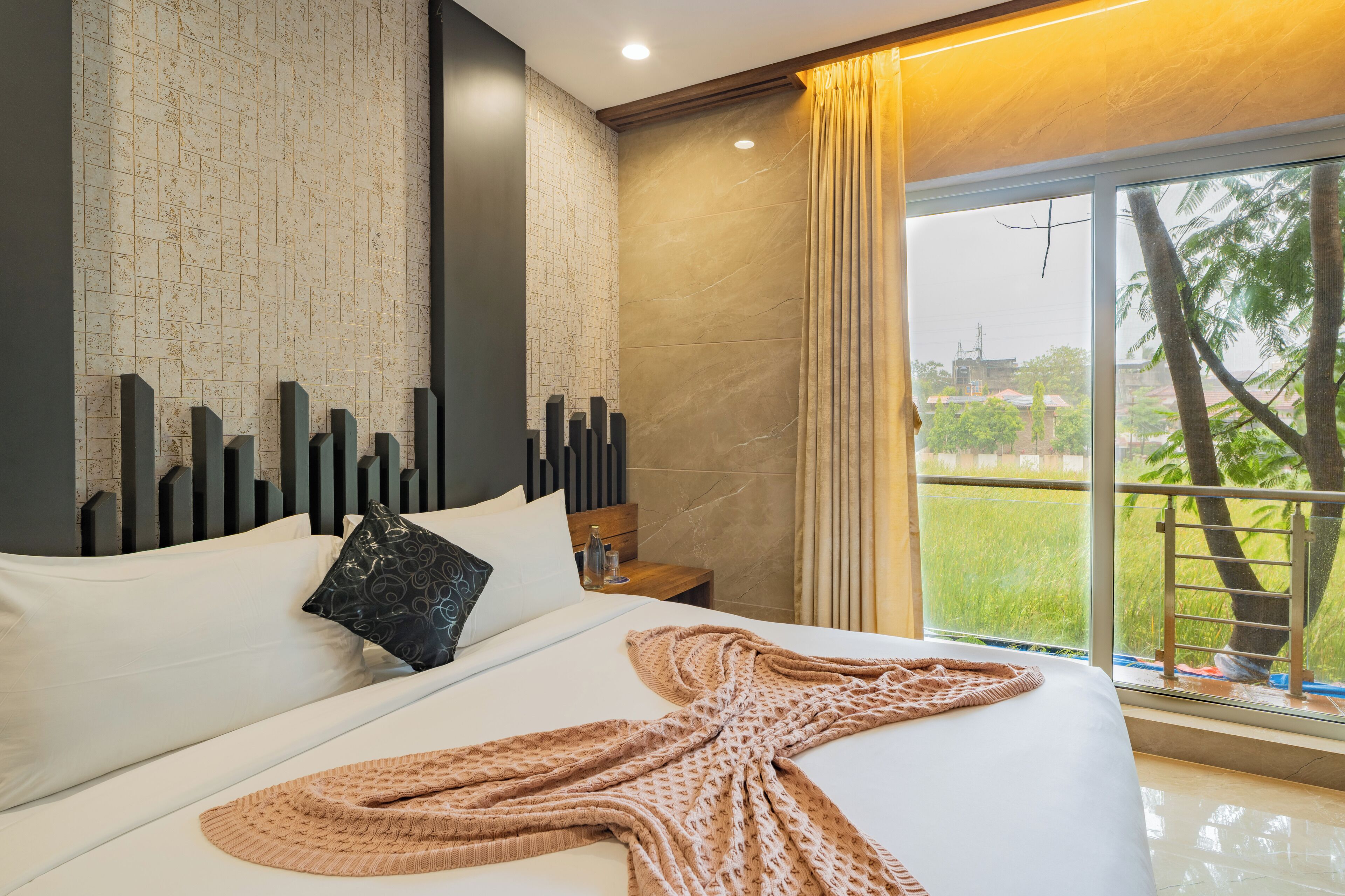 A modern hotel room with a large bed, a view of greenery outside, and a cityscape visible through the window.