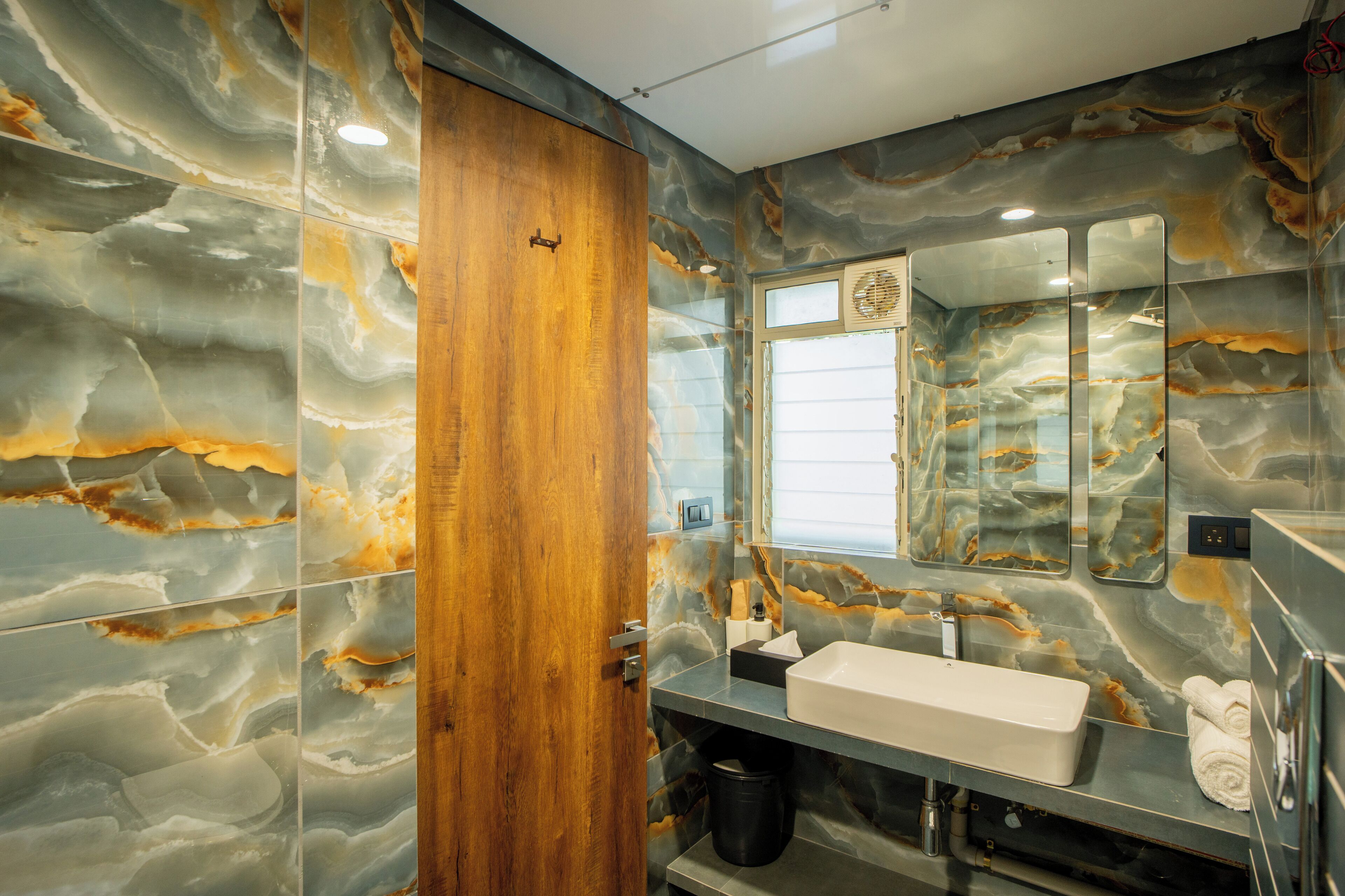 A bathroom with a marble wall, a wooden door, a sink, and a mirror.