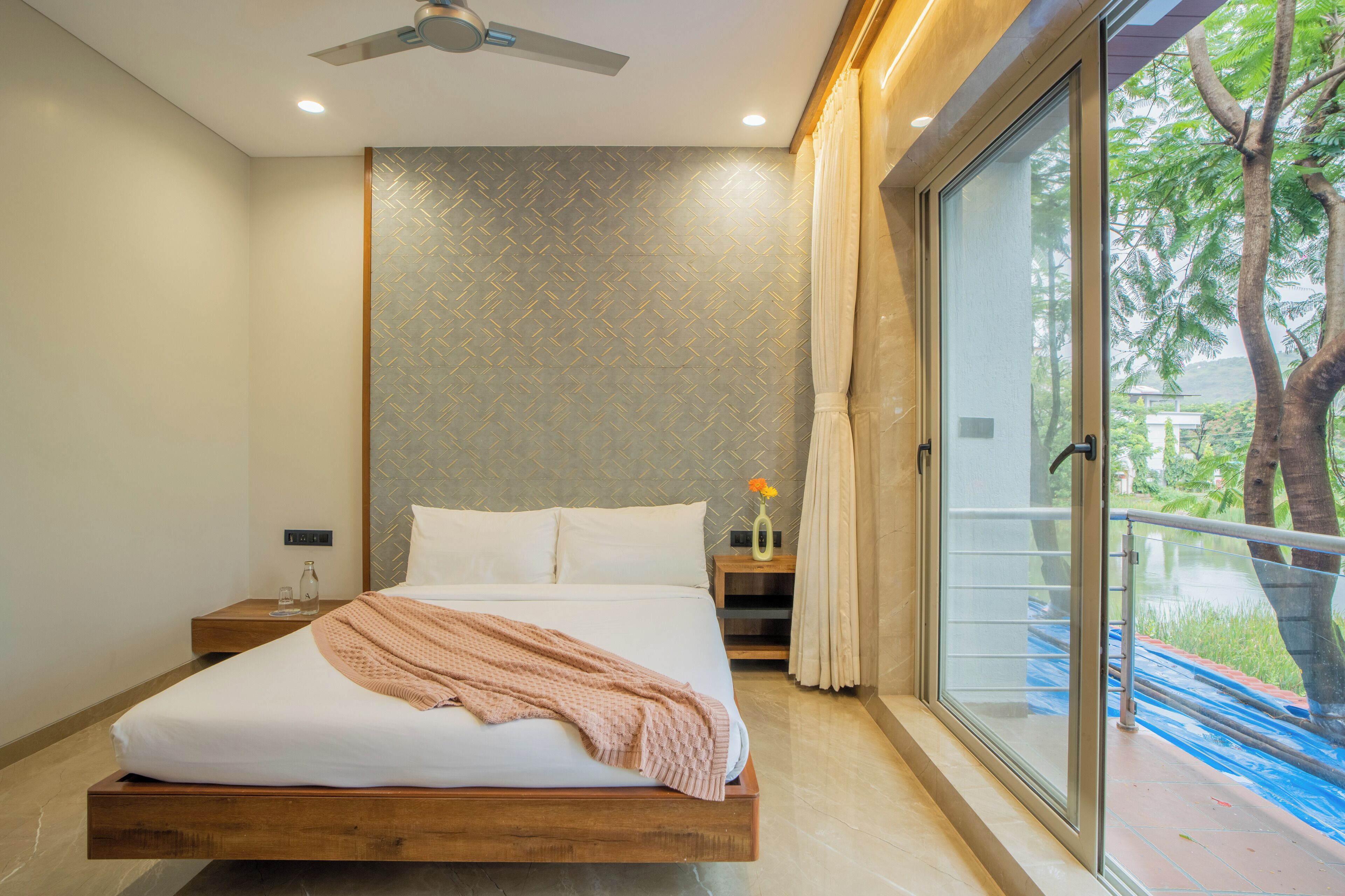 A modern bedroom with a large bed, a nightstand, a vase with flowers, and a view of a tree through a glass door.