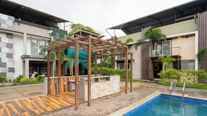 Un área de piscina con un pérgola de madera, un rincón con cascada y una piscina con borde de azulejos.