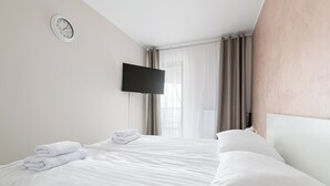 Ett hotellrum med en säng, en platt-TV, en väckarklocka och ett fönster med gardiner.