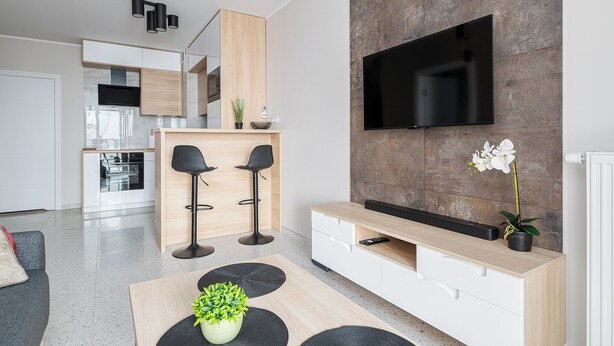 Ett modernt vardagsrum med en platt-TV som är monterad på en vägg av sten, ett matbord i trä, två svarta barstolar och ett köksområde i bakgrunden.