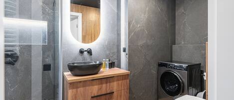 A modern bathroom with a large oval mirror, wooden vanity, black sink, and a built-in washing machine.