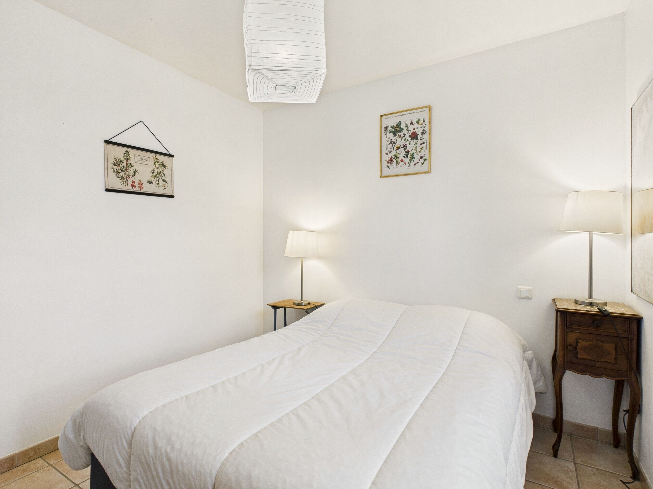 Une chambre à coucher comprenant un lit, deux tables de chevet, un luminaire suspendu et deux reproductions encadrées de motifs botaniques accrochées au mur.