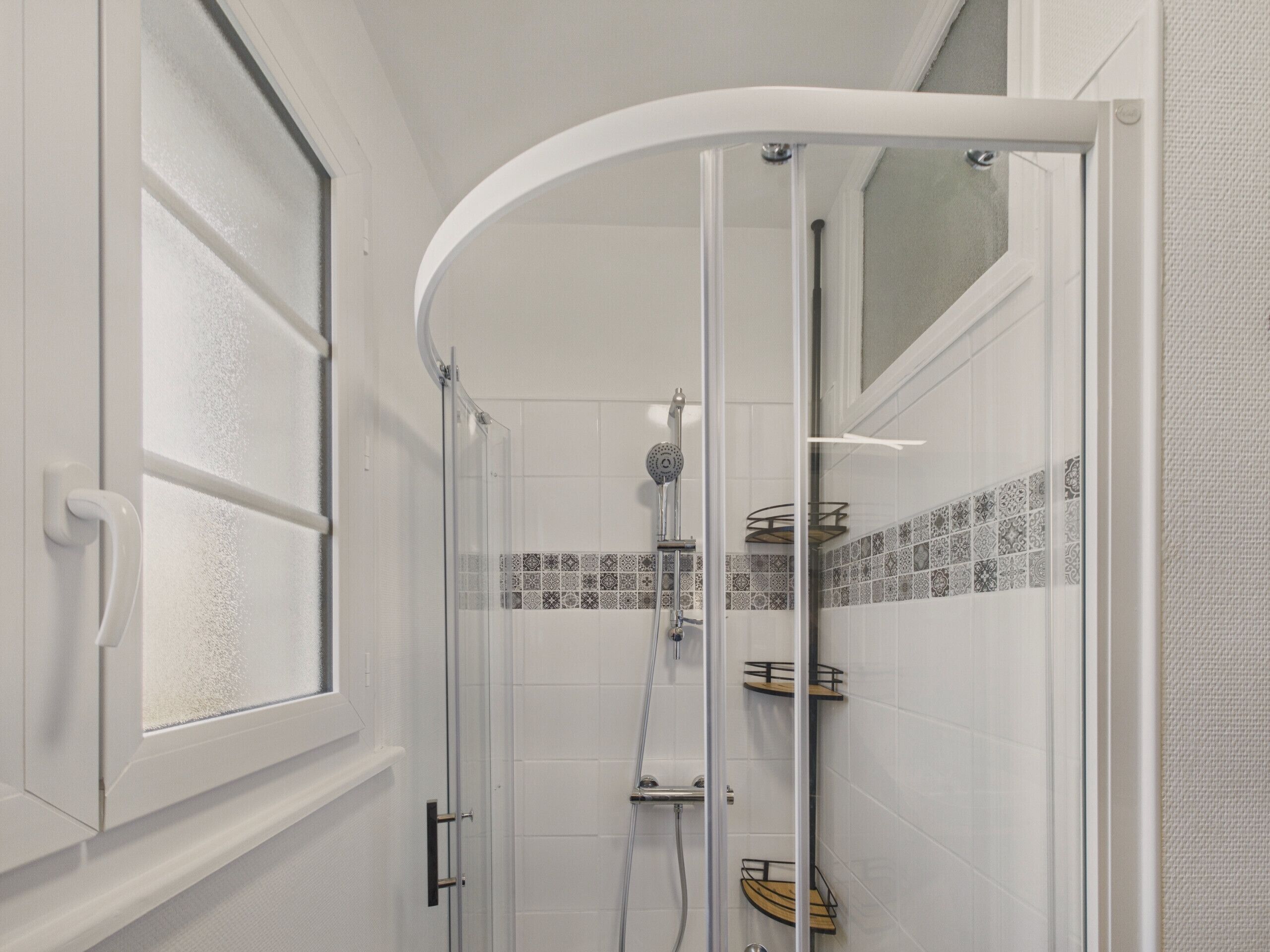 Et moderne badeværelse med en glasafskærmet brusekabine, hvide flisevægge og et vindue med matteret glas.