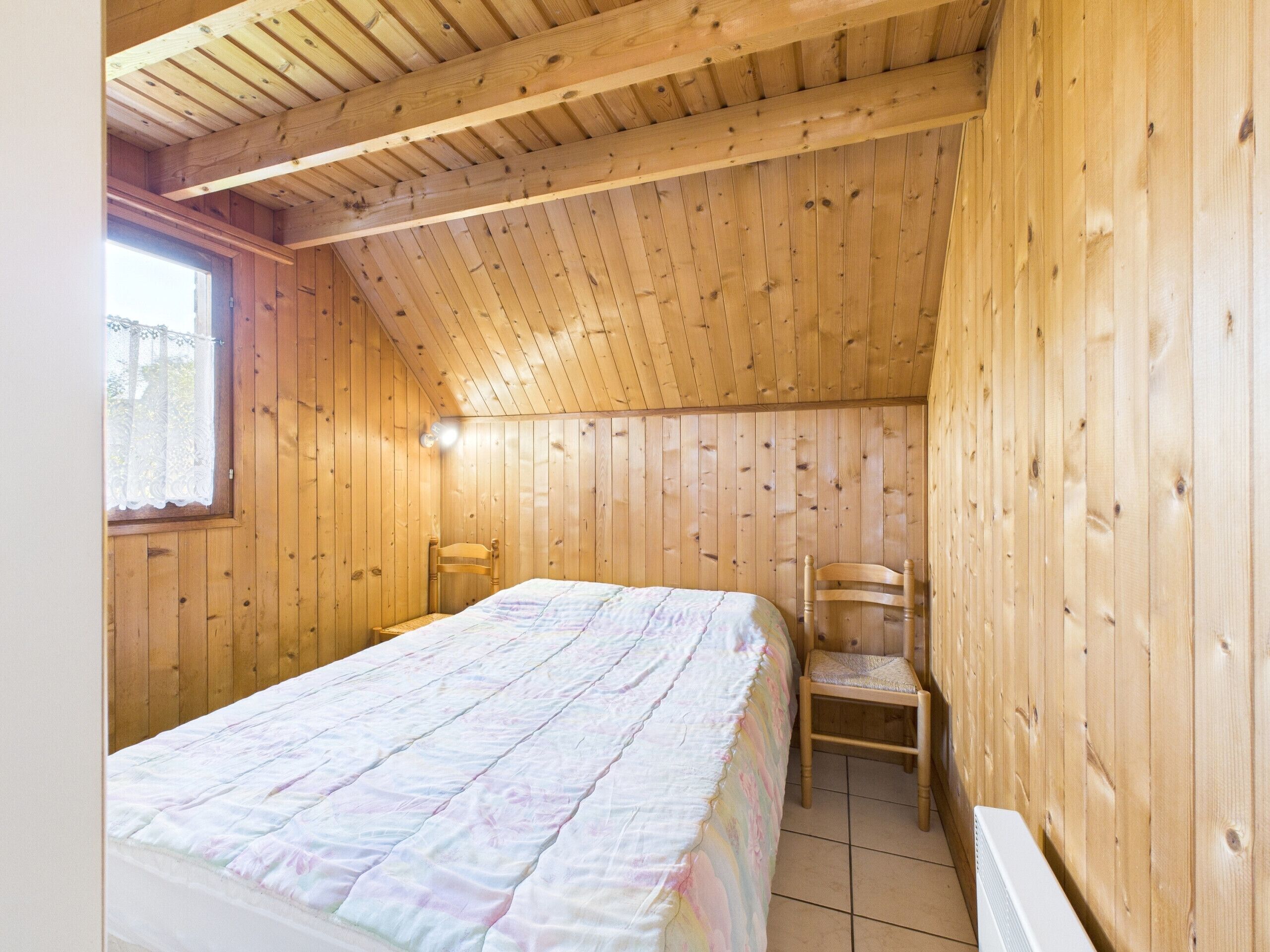 A bedroom with wooden walls and ceiling, a bed with a colorful quilt, a chair, and a window with a curtain.