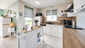 Une cuisine moderne avec des armoires blanches, un plan de travail en bois et un petit îlot central. On y trouve deux fenêtres avec des stores verts, un luminaire au plafond et divers appareils de cuisine.