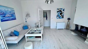 Un salon avec un canapé blanc, une table basse en verre et un meuble blanc. Il y a un tableau au mur et une lampe suspendue au plafond.