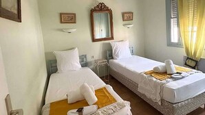 Une chambre d’hôtel avec deux lits, chacun recouvert de draps blancs et d’une couverture jaune. On y trouve deux lampes fixées au mur, un miroir et une fenêtre avec des rideaux jaunes.