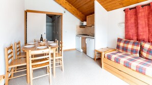 Een compacte kamer met een houten eettafel en stoelen, een kitchenette met een magnetron, en een bank met geruit kussens.