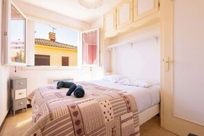 Un dormitorio con cama, una ventana con vista a edificios, una mesita de noche y un armario.