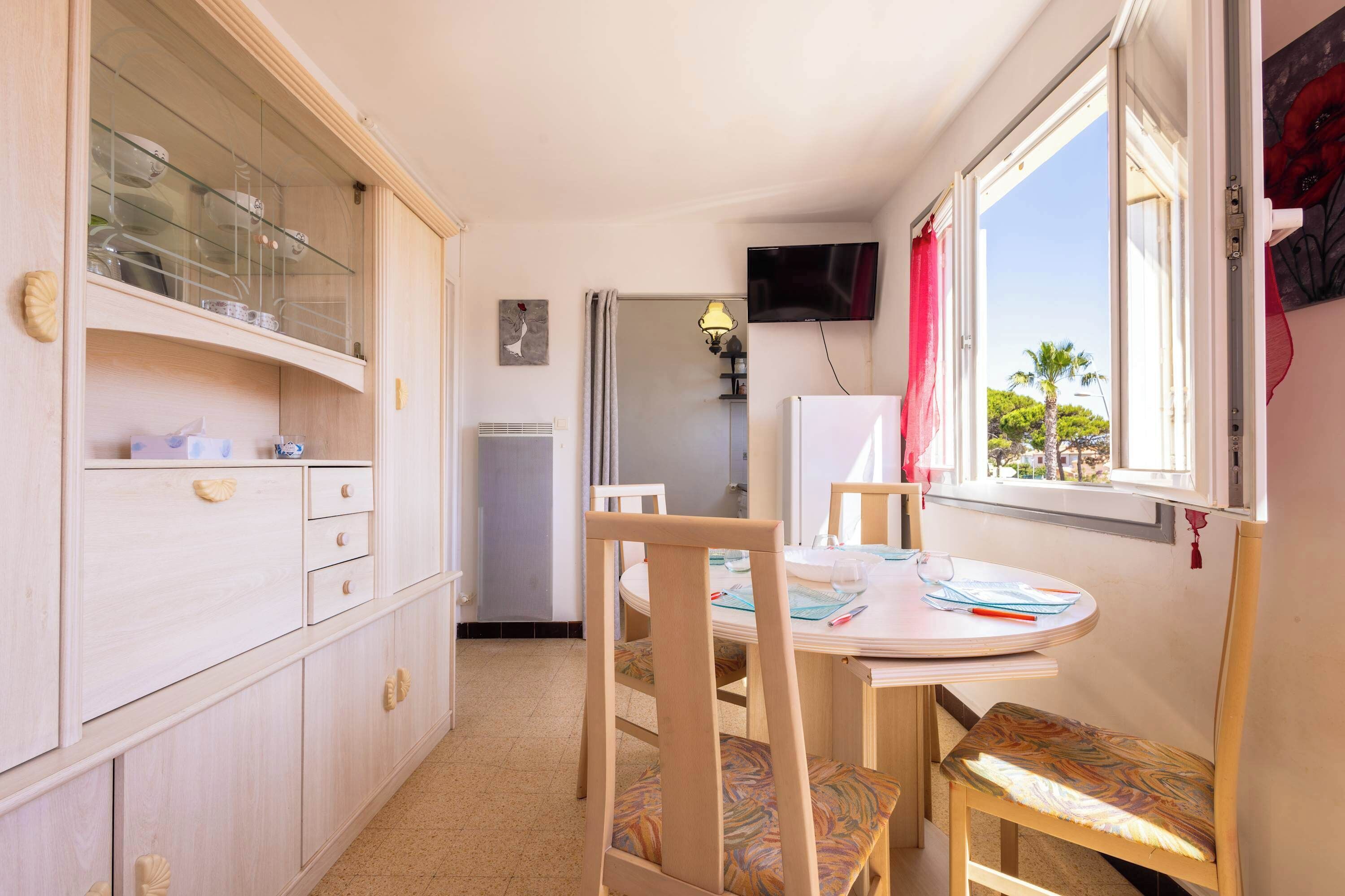 Una cocina compacta con zona de comedor, un televisor montado en la pared y un ventanal con vistas a palmeras.