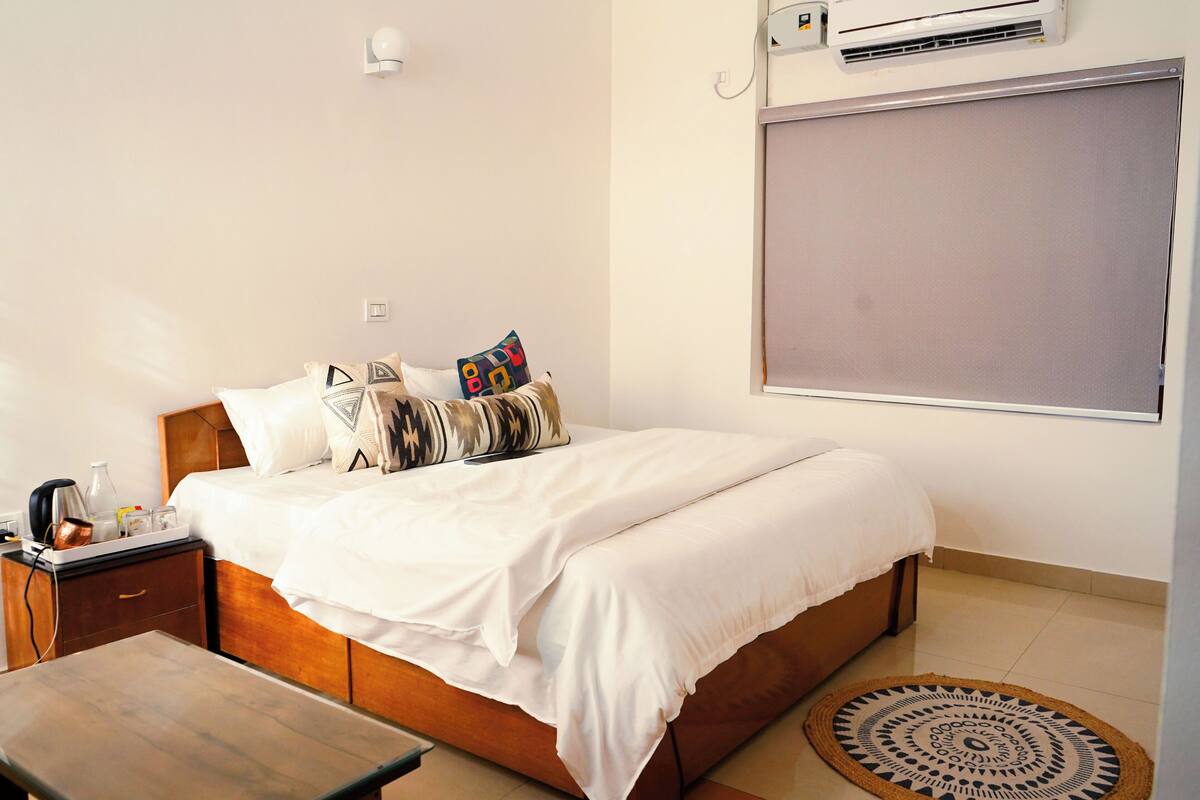 A bedroom with a bed, bedside table, and a window with an air conditioning unit.