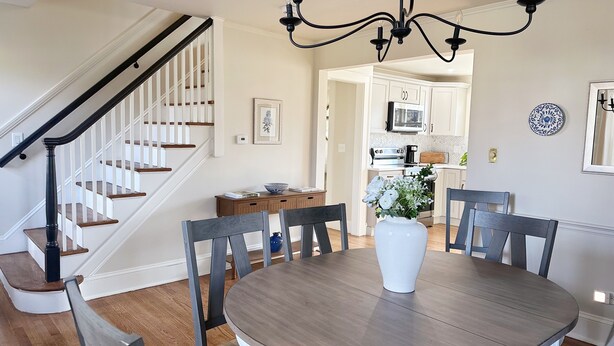 A dining area with a round table, four chairs, a vase with flowers, a staircase with white railings, and a kitchen in the background. - The Majestic View / Luxury Stay Gulf Course Views / 4-Bedroom House (Bloomfield)