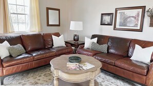 A living room with a brown leather sectional sofa, a round coffee table, and a lamp. There are framed pictures on the wall and a window with curtains. - The Majestic View / Luxury Stay Gulf Course Views / 4-Bedroom House (Bloomfield)