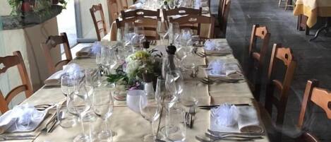 Een lange eettafel, gedekt met witte tafelkleden, glazen en houten stoelen, in een restaurant met gewelfde plafonds en potplanten.