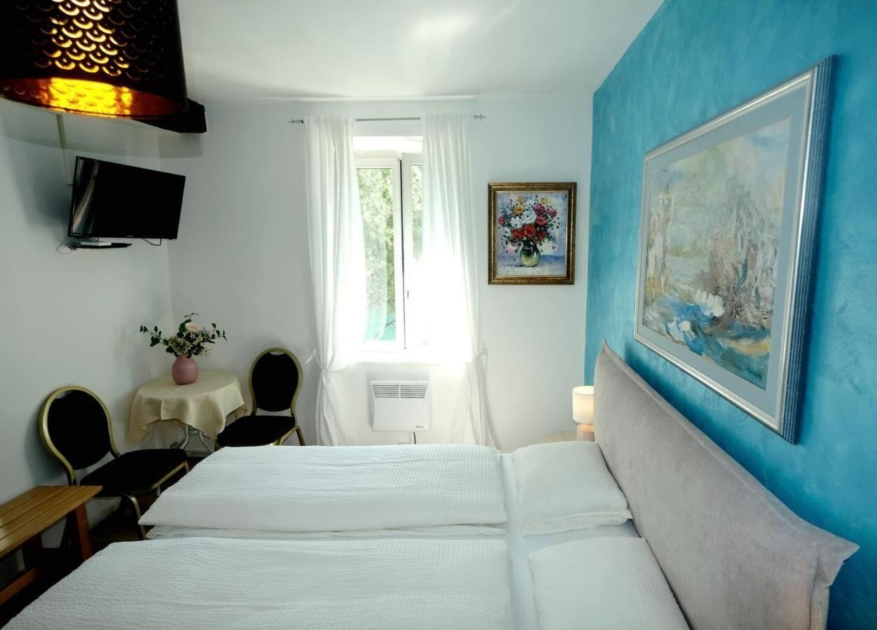 A hotel room with a bed, a desk, a chair, a TV, a window with curtains, and a painting on the wall.