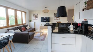 A modern kitchen with a black pendant light, a dining area with a white table and chairs, and a living room with a sofa and a TV. - LE PETIT SESAME L' évasions cosy jacuzzi terrace wifi parking 2 steps stadiumDF (Saint-Denis)
