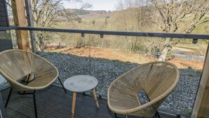 Une terrasse en bois avec deux fauteuils en osier et une petite table ronde, d’où l’on peut admirer un paysage rural avec des arbres et des collines.