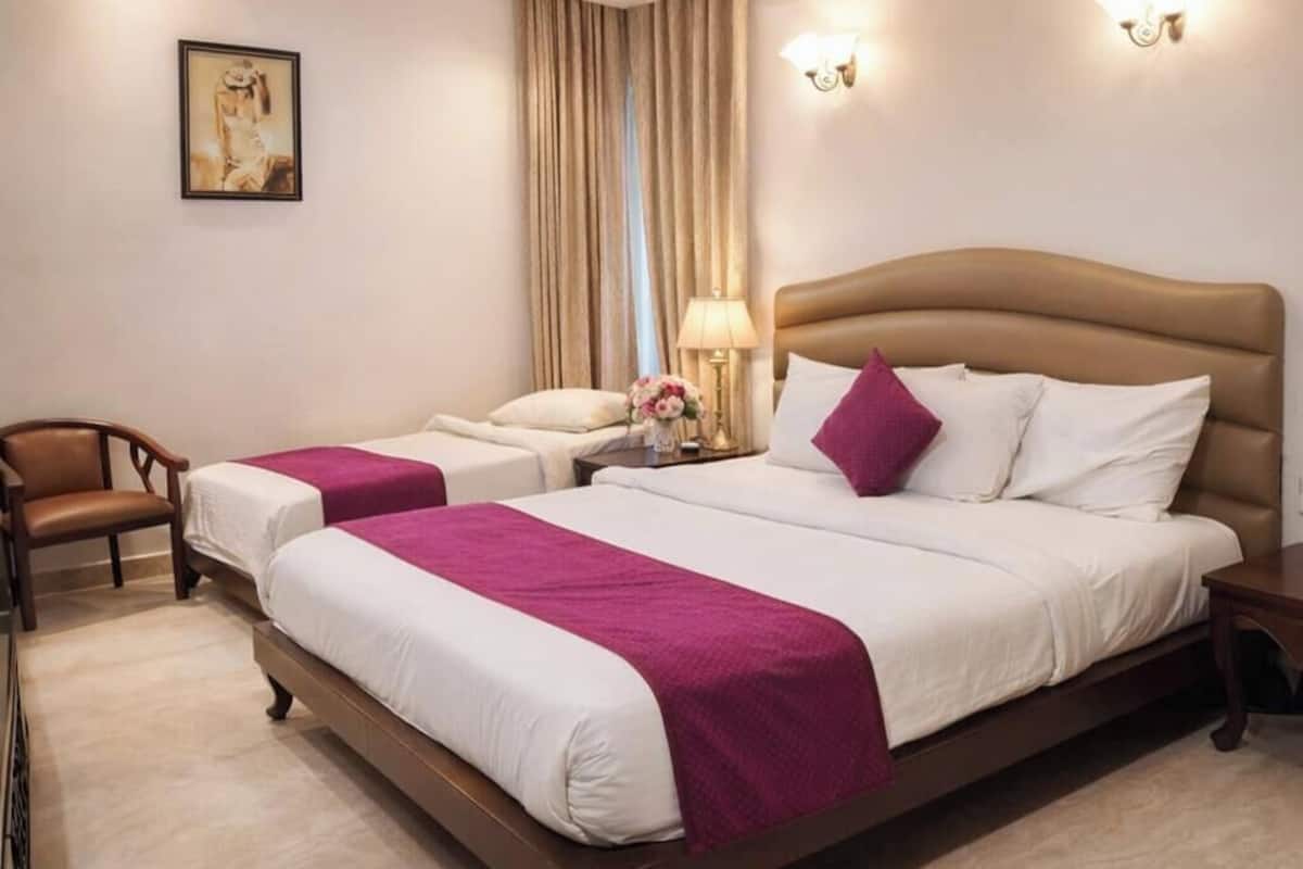 Habitación de hotel con una cama grande, dos camas más pequeñas, una silla, una mesita de noche con lámpara y un cuadro en la pared.