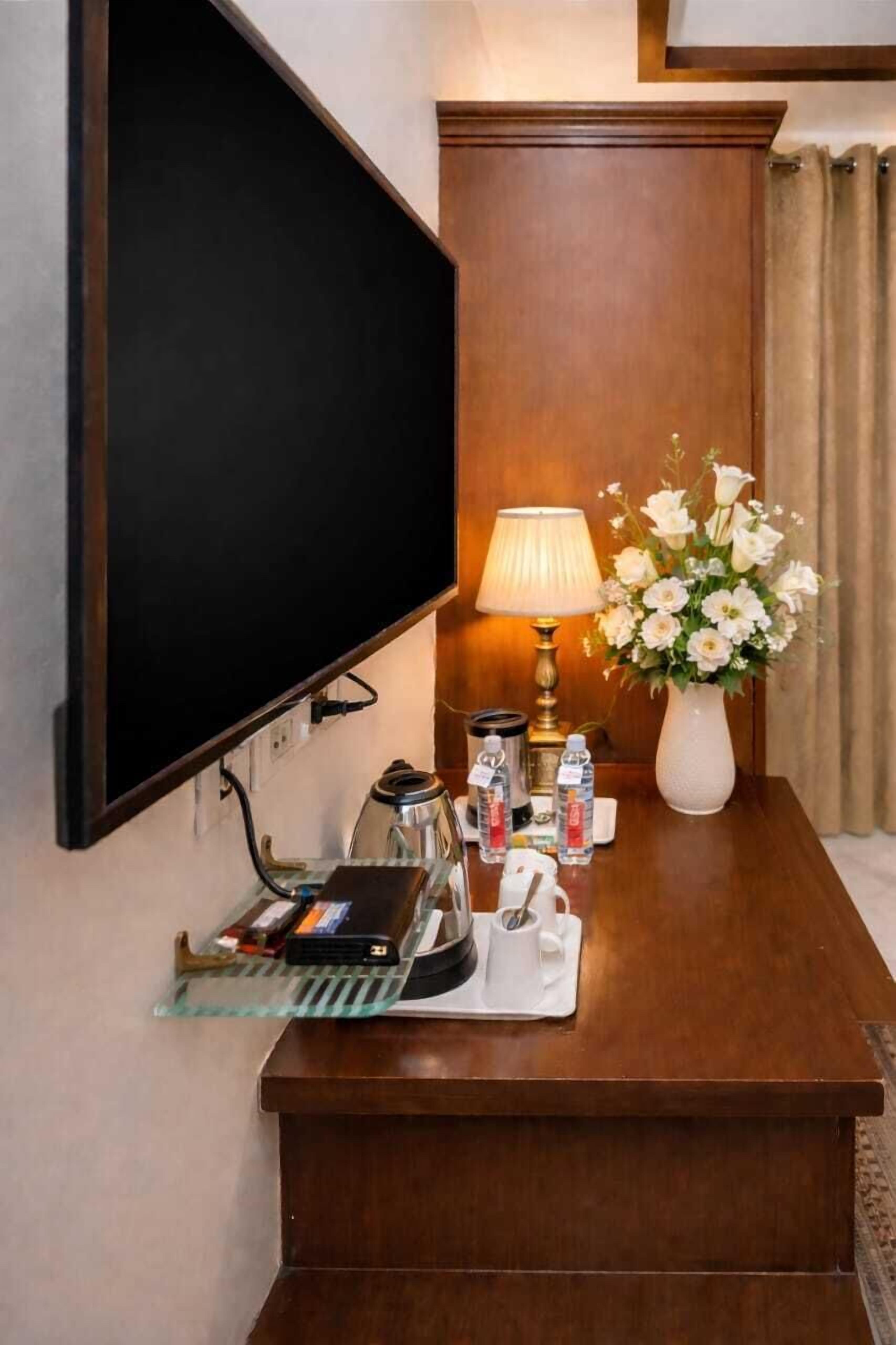 A hotel room with a flat-screen TV mounted on the wall, a wooden desk with a lamp, a vase of flowers, and various toiletries.