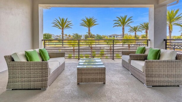 Een balkon met rieten meubels, een tafel met glazen blad en uitzicht op palmbomen en huizen.