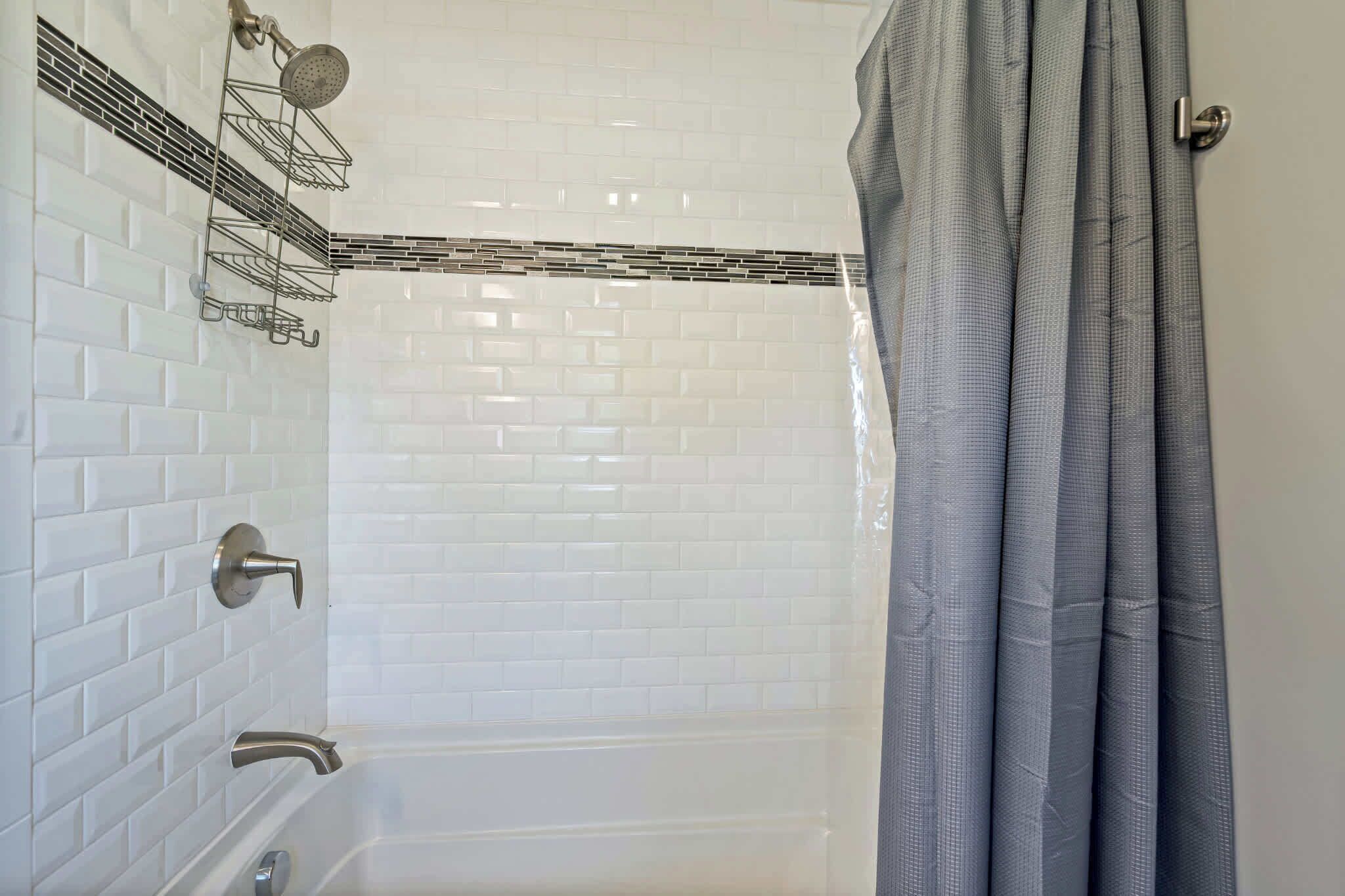 Ein Badezimmer mit weißen Fliesenwänden, einer Badewanne, einer Dusche mit Duschbrausehandstück und einem grauen Duschvorhang.
