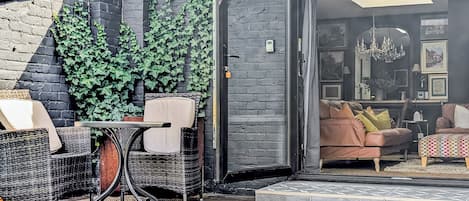 En åben terrasse med fletmøbler, en murstensvæg og udsigt til en stue med sofa og puder.