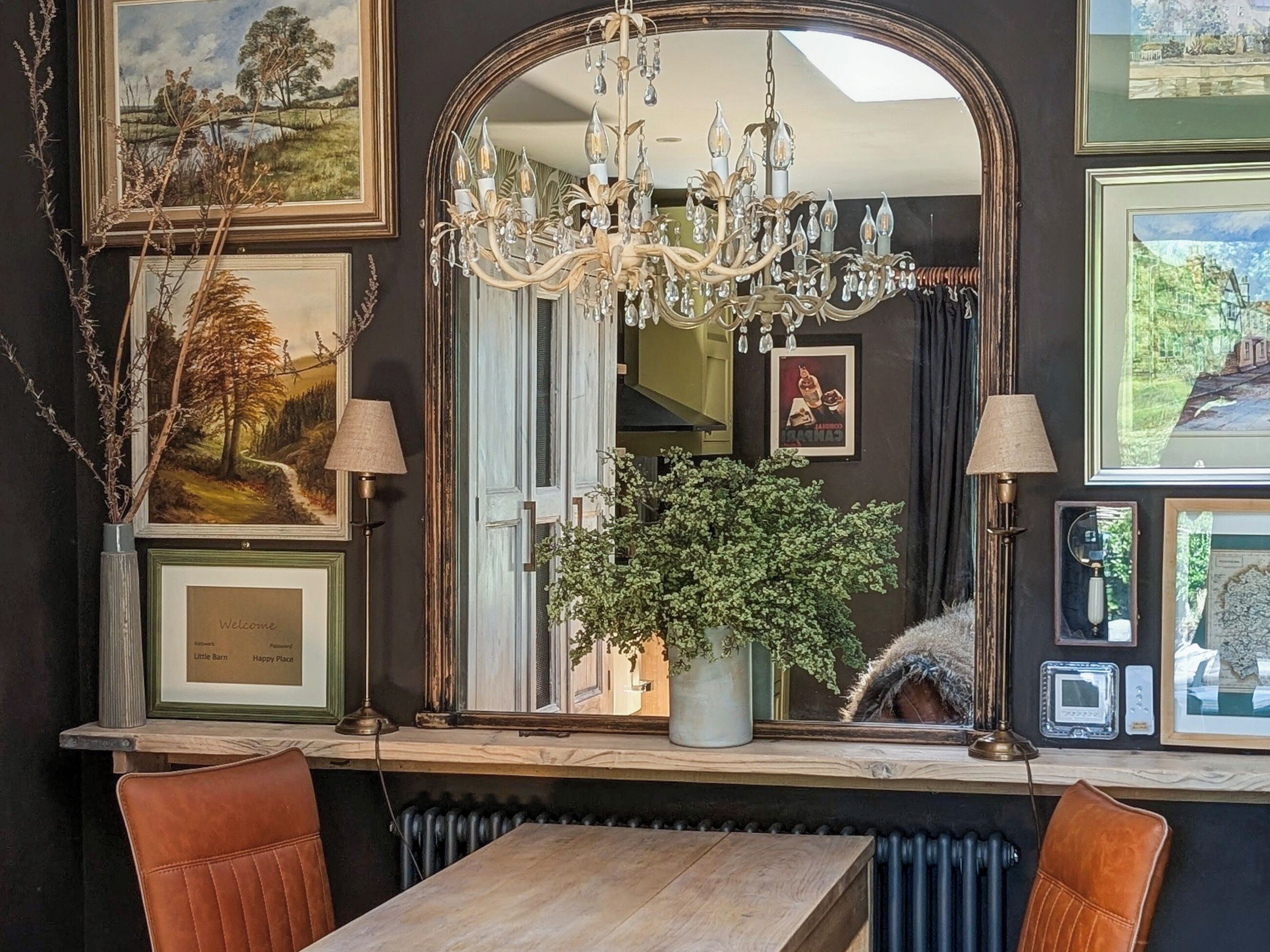 A room with a large mirror, a chandelier, framed artwork, a potted plant, and a wooden table.