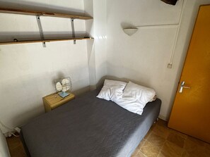 Uma cama de solteiro com almofadas brancas, uma mesa de cabeceira com luminária e uma prateleira de madeira acima.