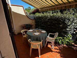 A patio with a round table set for four, surrounded by chairs, under a striped awning. - Charming pavilion in Argelès-sur-Mer - Beach 5 min, air conditioning, pets allowed (Argelès-sur-Mer)
