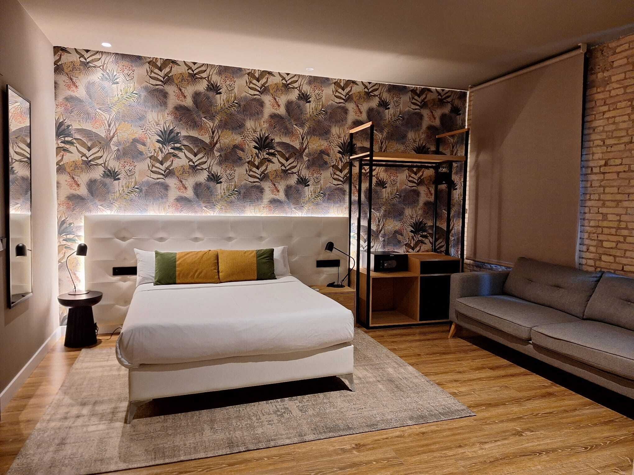 A modern bedroom with a large bed, a grey sofa, a wooden floor, and a patterned wallpaper.